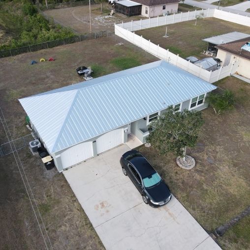 An aerial view of a light-colored single-story house with a metal roof and a black car parked in the concrete driveway.