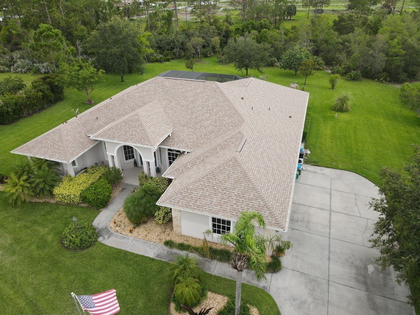 An aerial view of a single-story house with a light-colored shingled roof, a paved driveway, and a large grassy lawn.