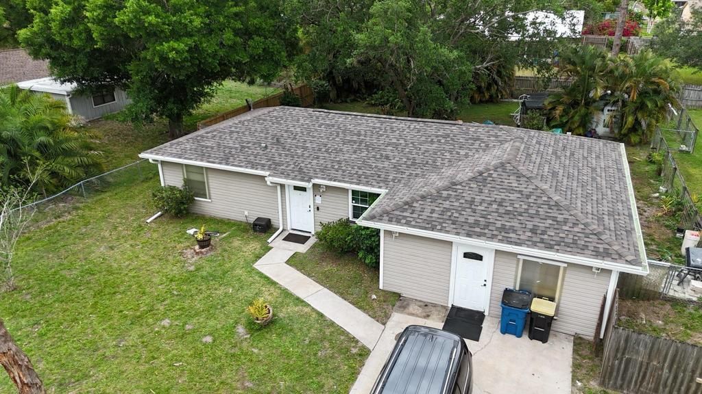 An aerial view of a single-story house with a light gray exterior and a shingled roof, set in a grassy, tree-filled yard.