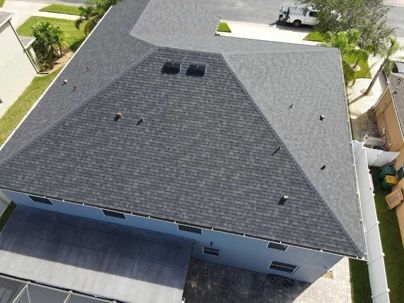 An aerial view of a blue house with a dark gray shingled roof, showing nearby properties and landscaping.