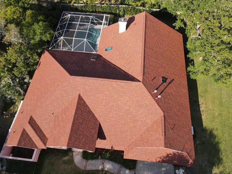 An aerial view of a residential house with a red shingled roof, a chimney, and a screened-in pool enclosure.