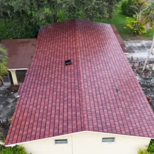 An aerial view of a house with a new red shingle roof and a light-colored exterior wall surrounded by trees and a driveway.