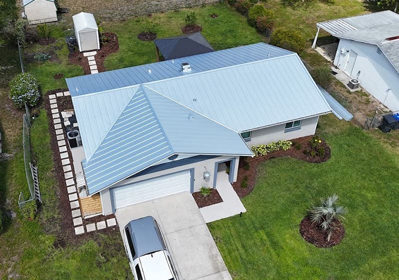 An aerial view of a single-story house with a light blue metal roof, a driveway with a car, and a large backyard.