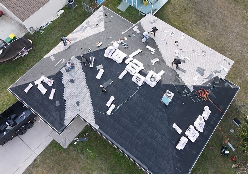 Aerial view of roofers installing new black asphalt shingles on a residential roof, with materials scattered across it.