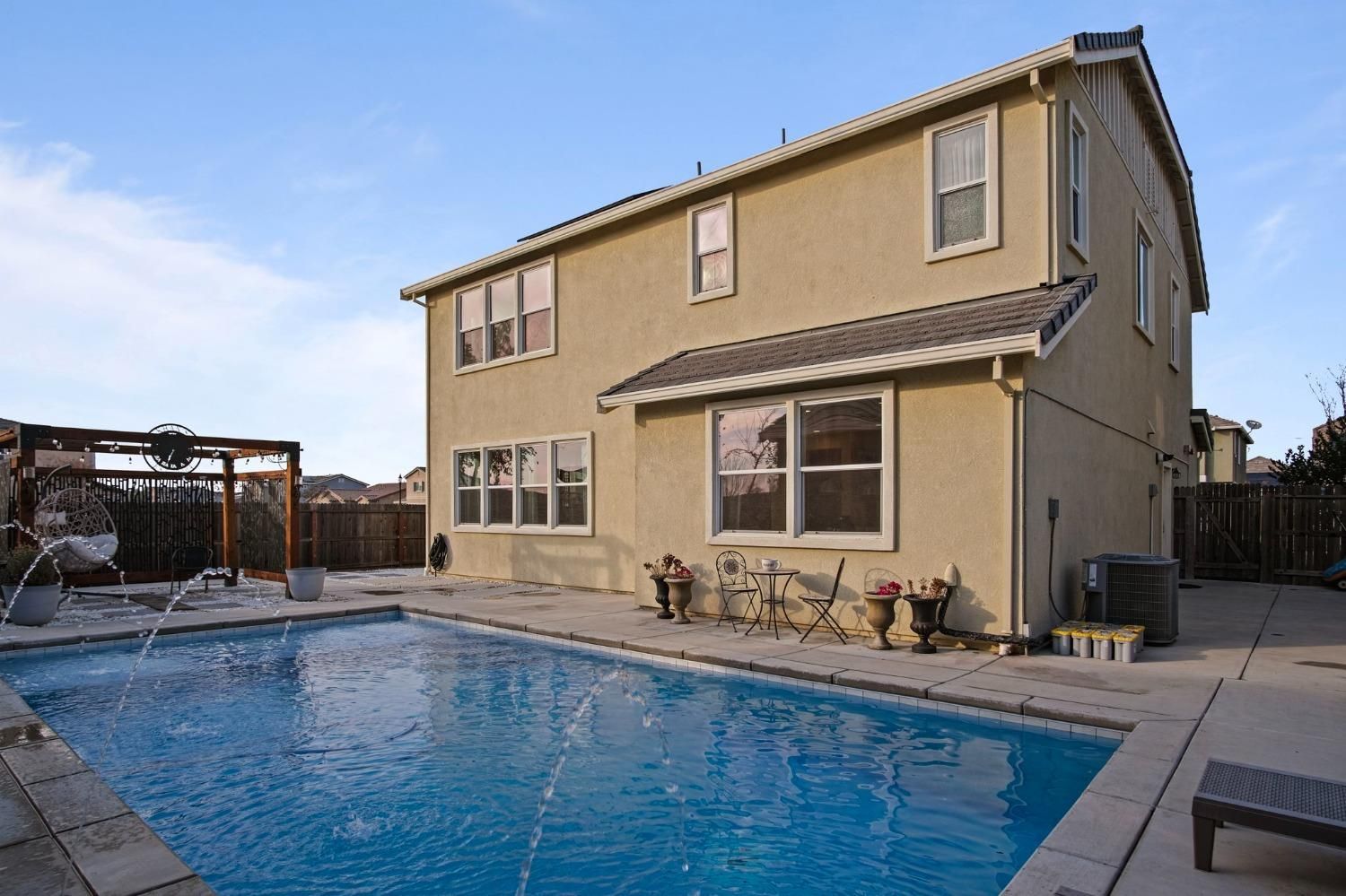 Two-story beige house with swimming pool, clear blue sky, concrete patio, and pergola.