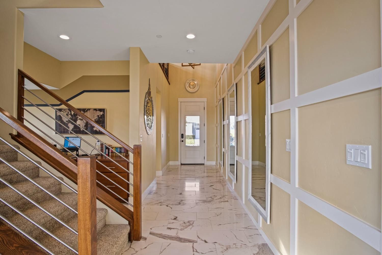 Hallway with staircase and decorative wall panels, beige walls, marble flooring, and front door at the end.