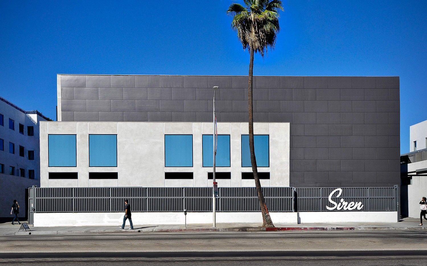 Modern building with gray facade, blue windows, palm tree, and a pedestrian walking on the street.