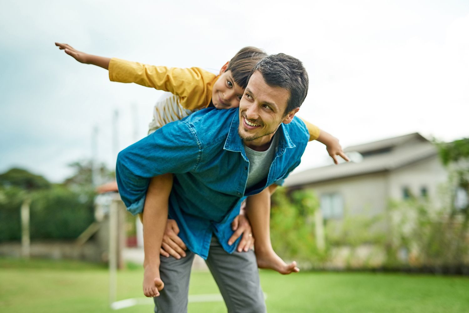 Adult carrying a child on shoulders in a backyard, both posing with outstretched arms. Adult carrying a child on shoulders in a backyard, both posing with outstretched arms.