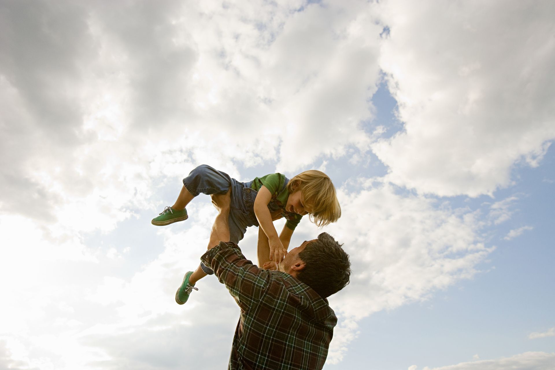 Man throwing a child up in the air against a cloudy sky. The child laughs, and the sun shines.