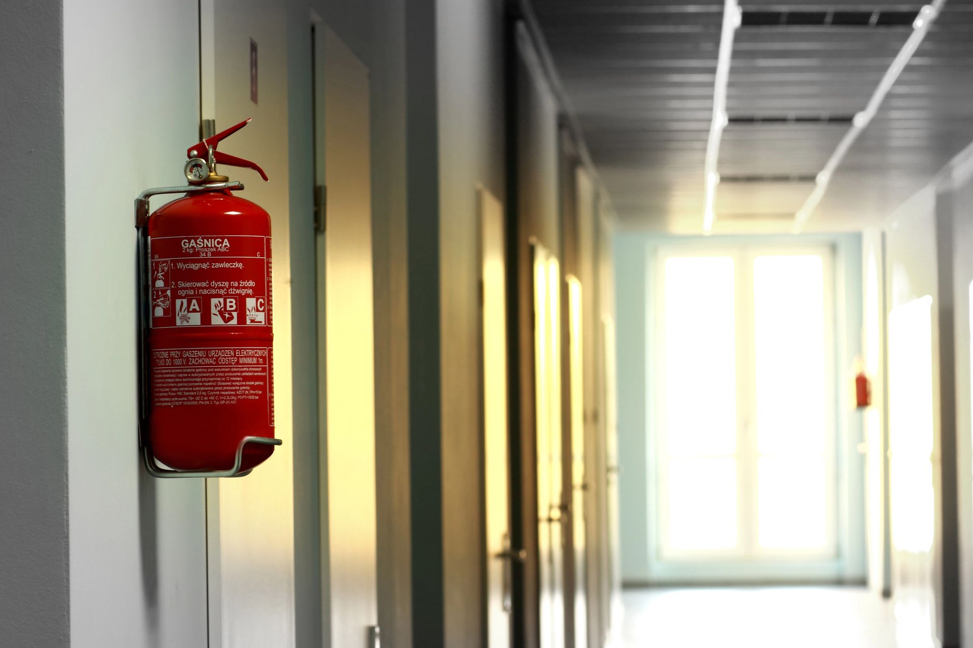 Extintor rojo montado en una pared gris en un pasillo con puertas cerradas.