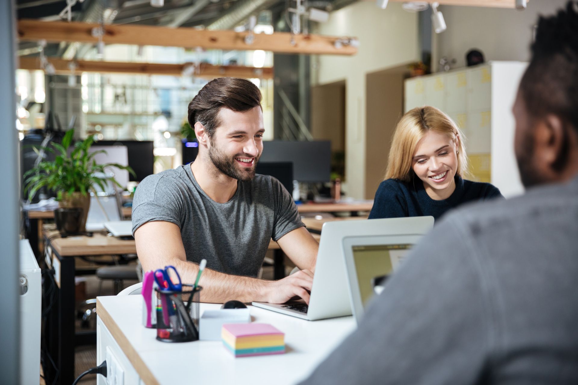 Deux collègues souriants, assis à un bureau dans un espace de travail lumineux, discutent avec une personne en face d'eux.