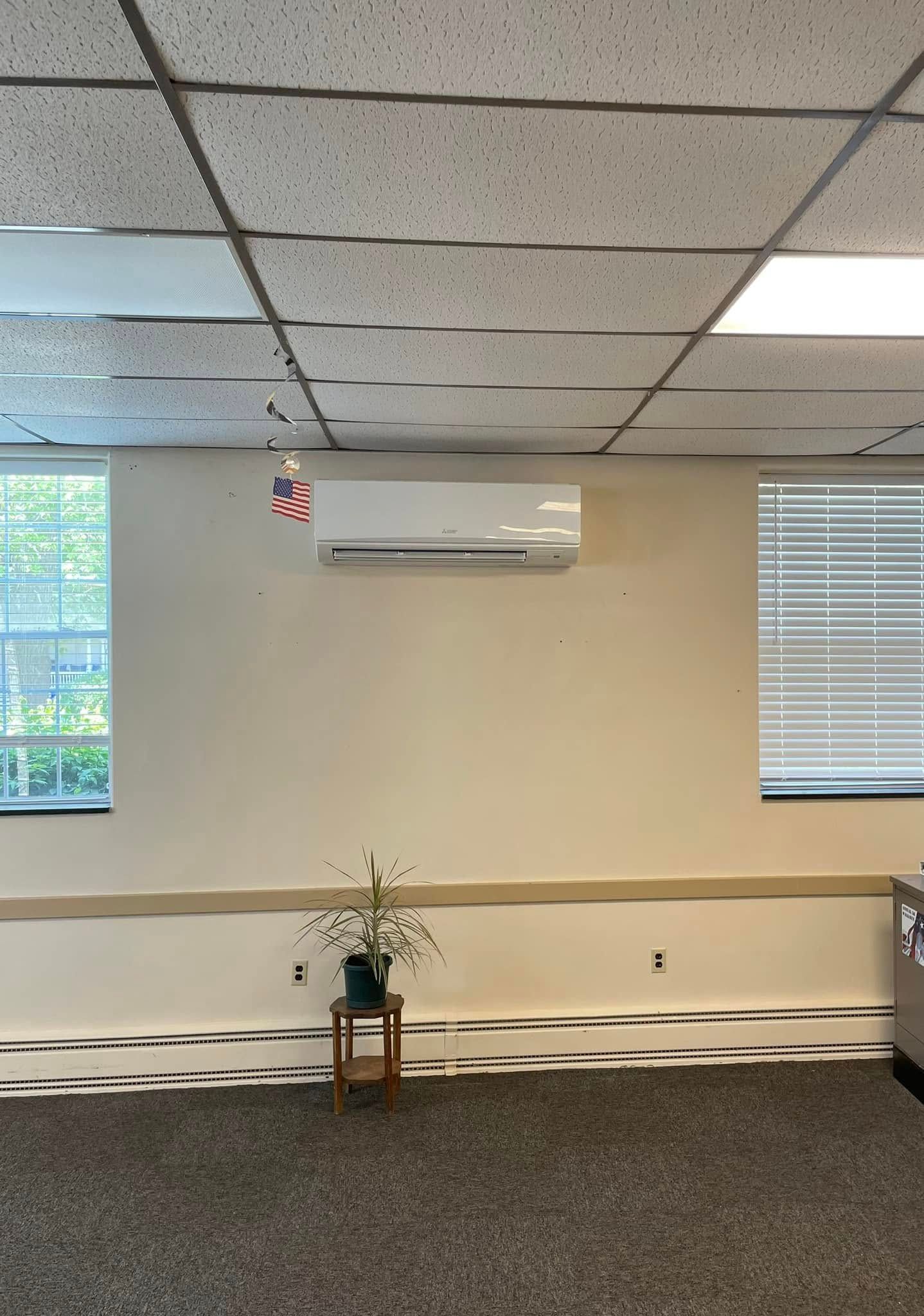 Empty office room with beige walls, drop ceiling, carpet, wall-mounted AC, and a small plant near the wall.