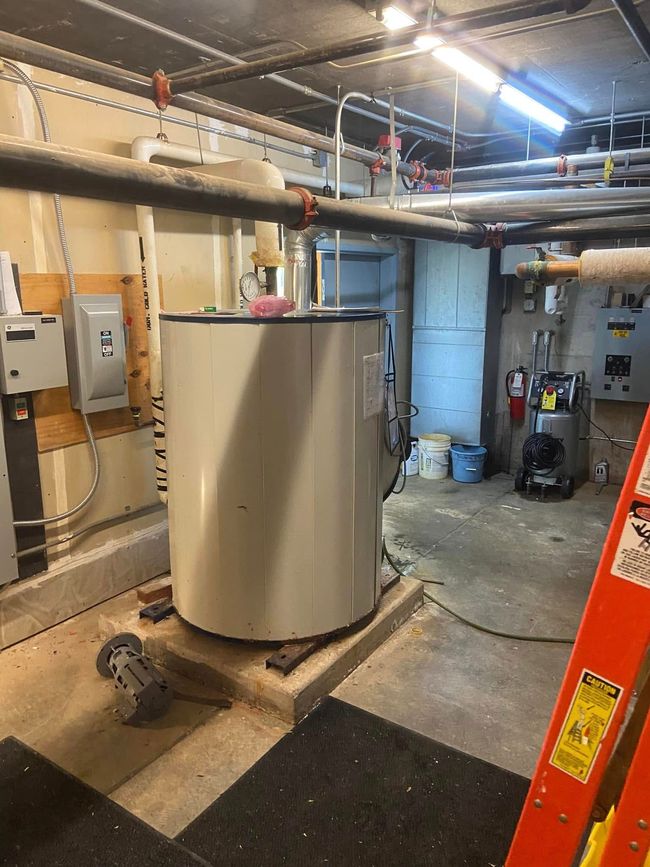 Utility room with a large water heater, exposed pipes, and a ladder in a basement-like workspace