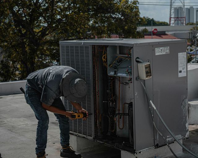 Worker fixing rooftop HVAC unit with a power tool on a building roof, equipment panels open.