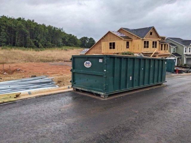 A green dumpster is parked on the side of the road in front of a house under construction.