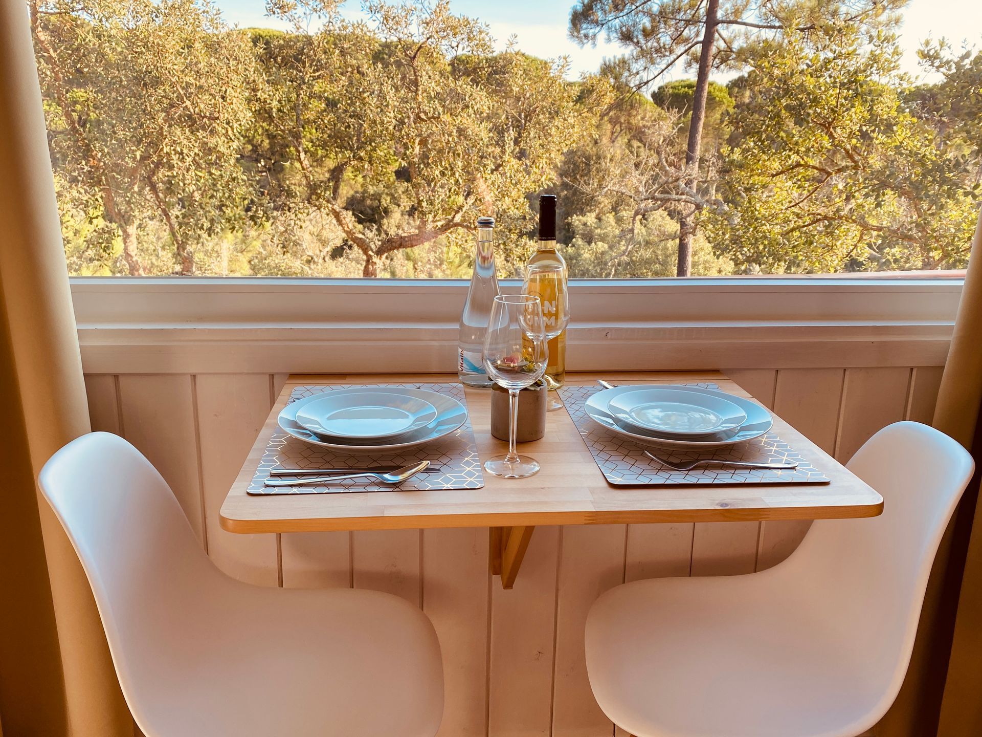 A small table with plates , glasses , and a bottle of wine in front of a window.