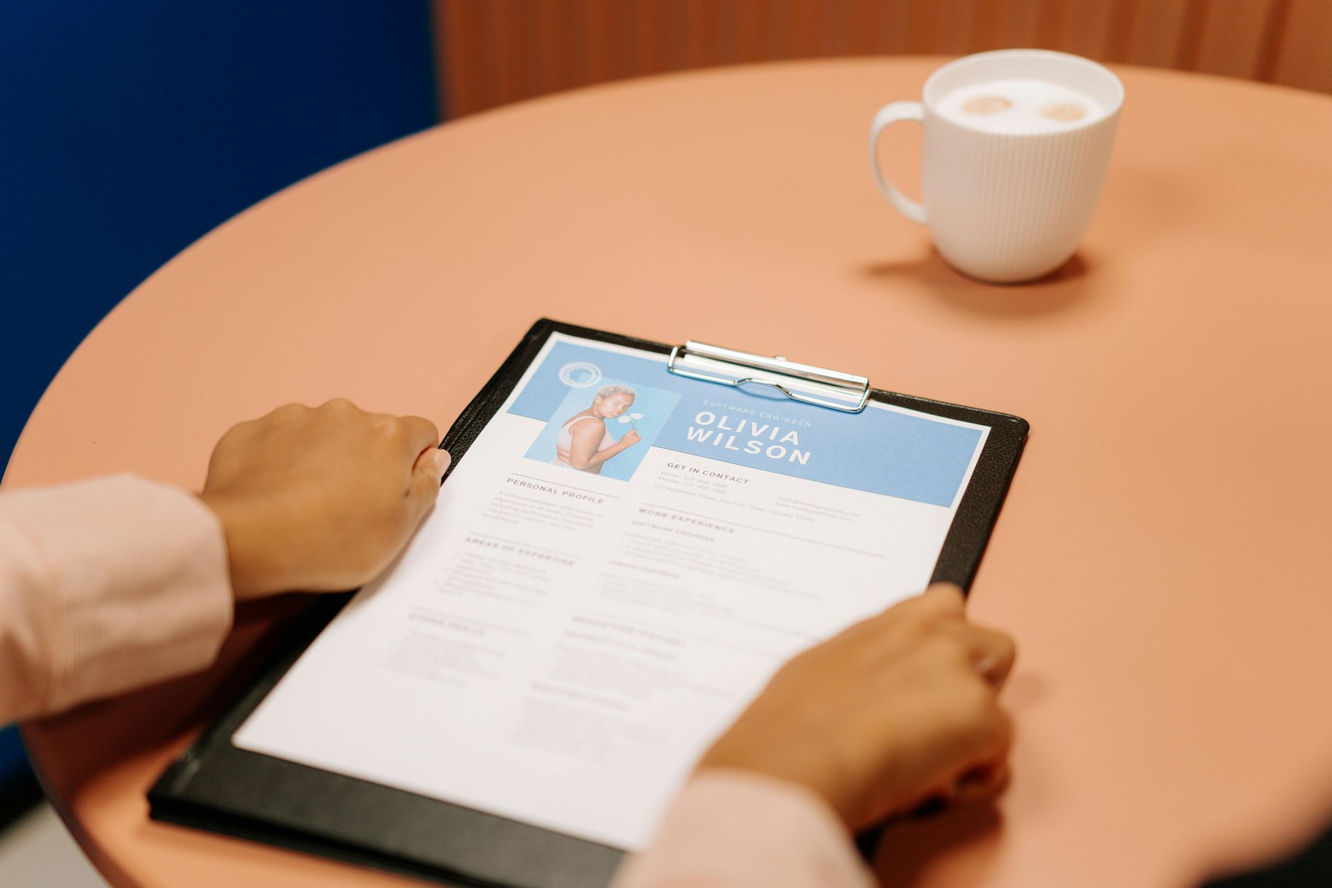 Person holding a resume on a clipboard, sitting at a round table with a mug.