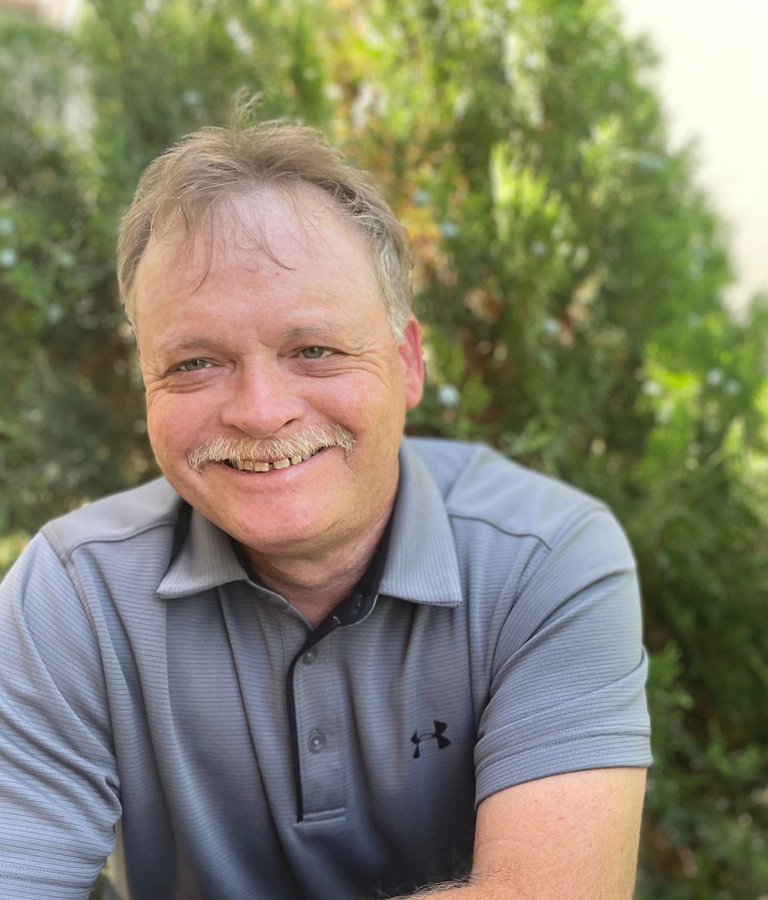 A man with a mustache is wearing a gray polo shirt and smiling.