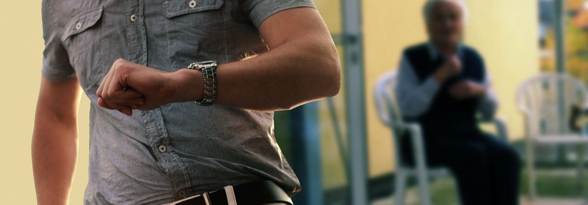 A man is looking at his watch while another man sits in a chair in the background.