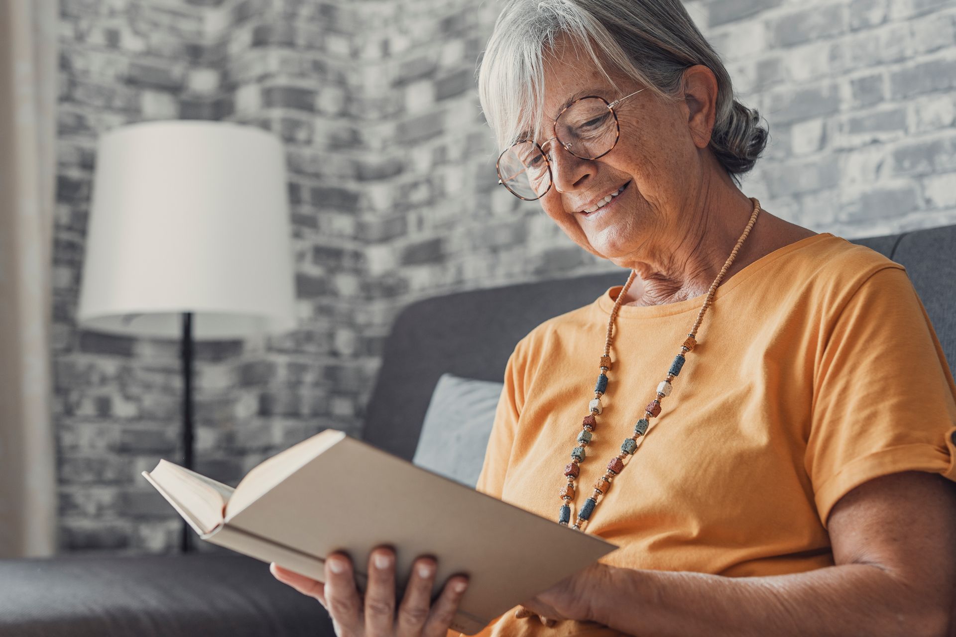 An elderly woman is sitting on a couch reading a book.