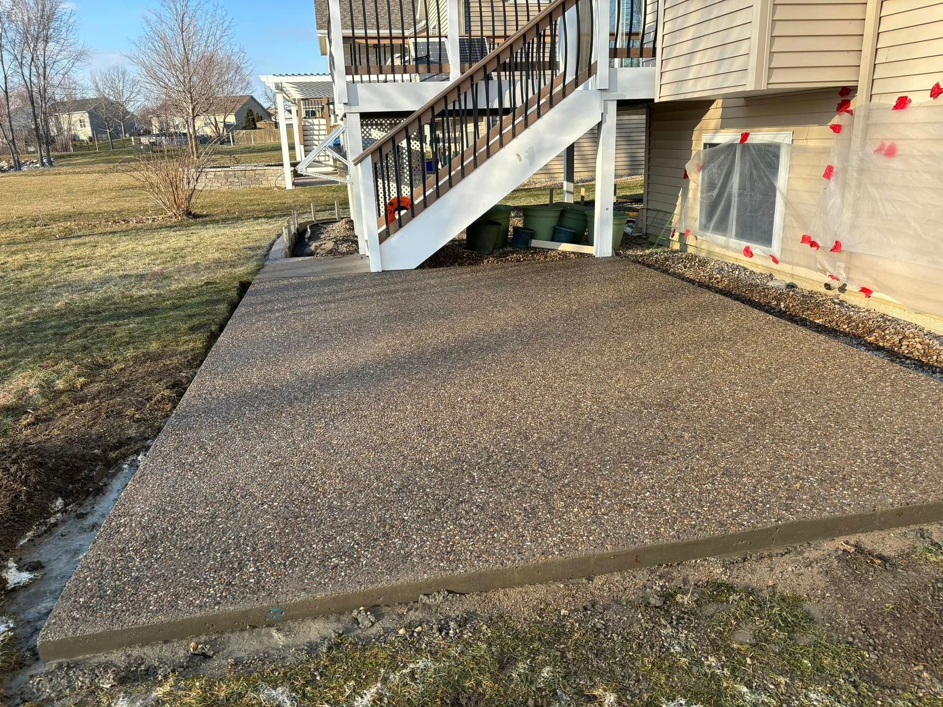 Concrete patio with exposed aggregate texture next to a house with a wooden deck and stairs.