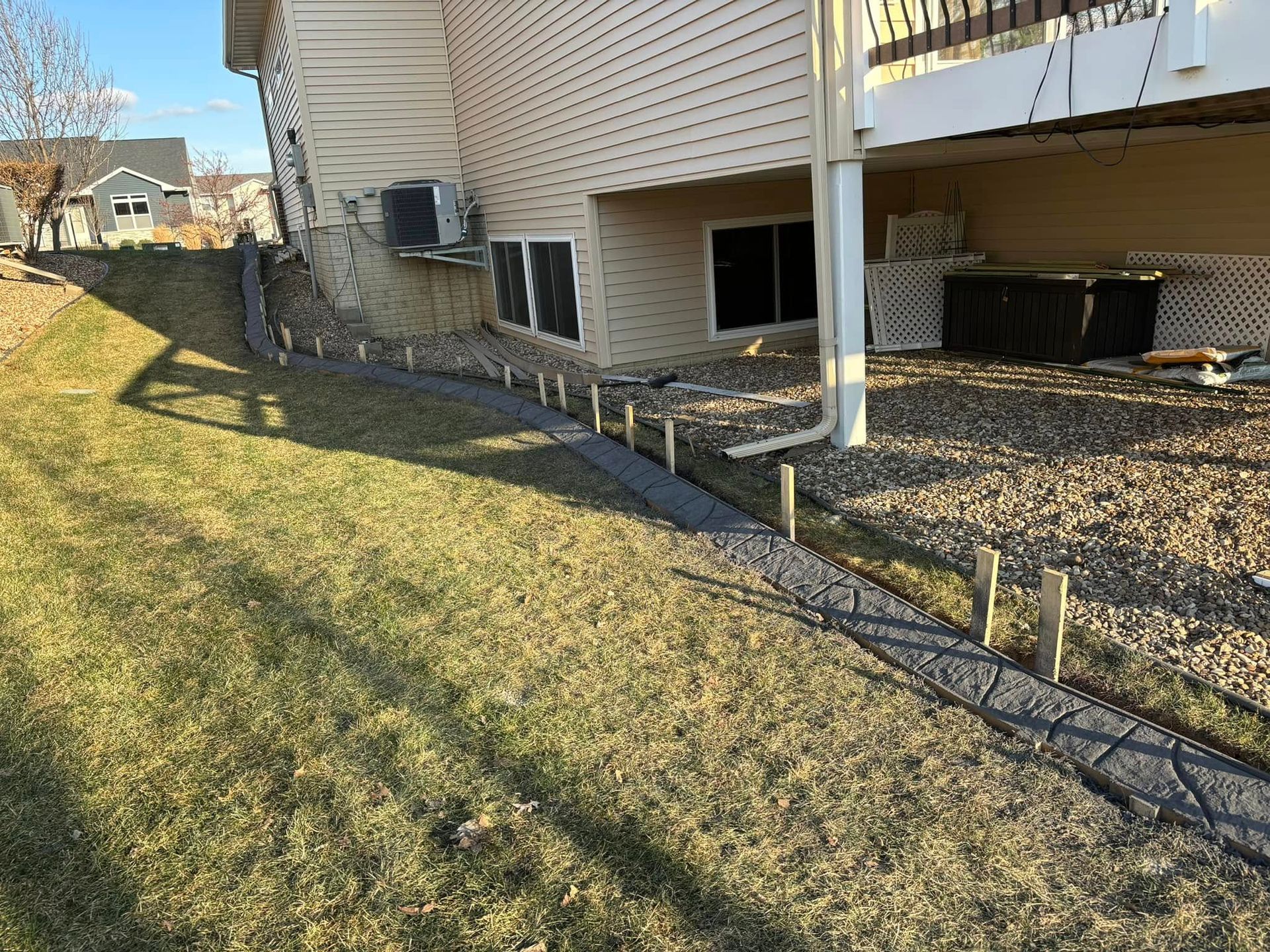A house with a retaining wall and sloped grassy yard.  Sunlit exterior with a wooden deck and a rock bed.