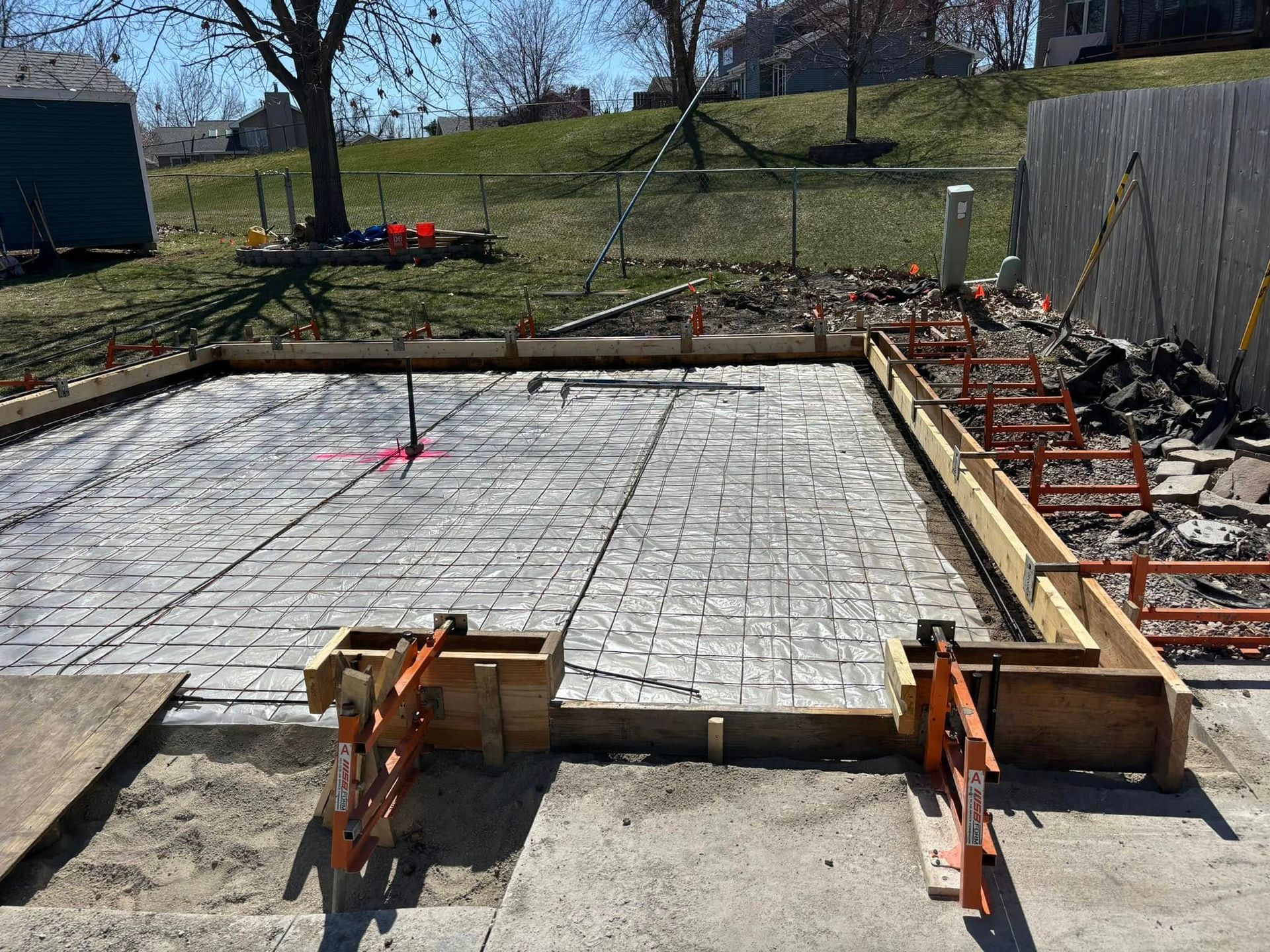 Concrete slab construction: wooden forms, rebar grid, orange bracing, residential backyard setting.