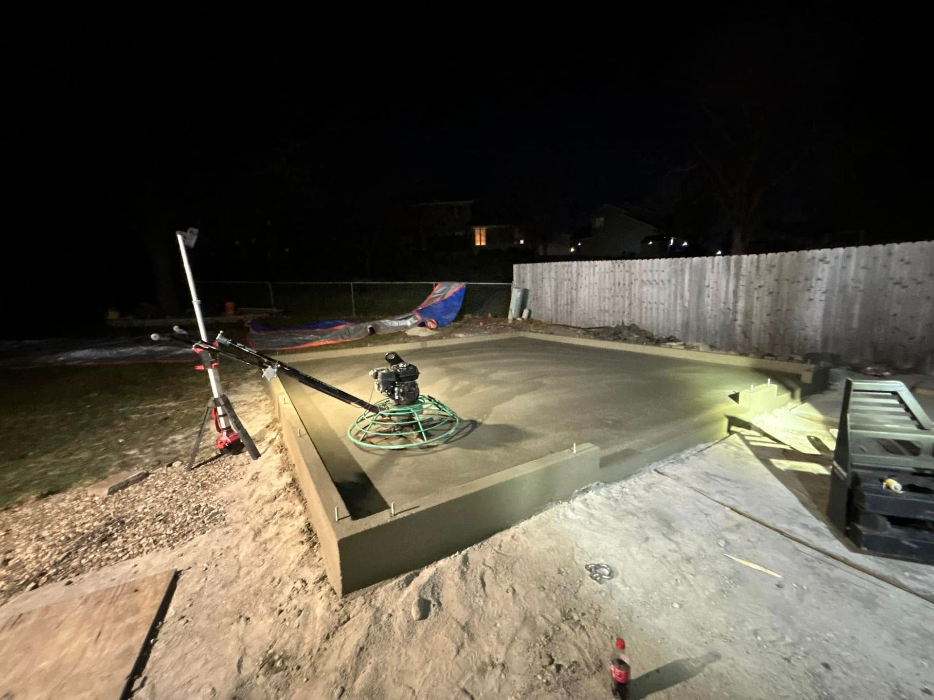 Nighttime view of a concrete slab being smoothed with a power trowel. Construction site lit by work lights.