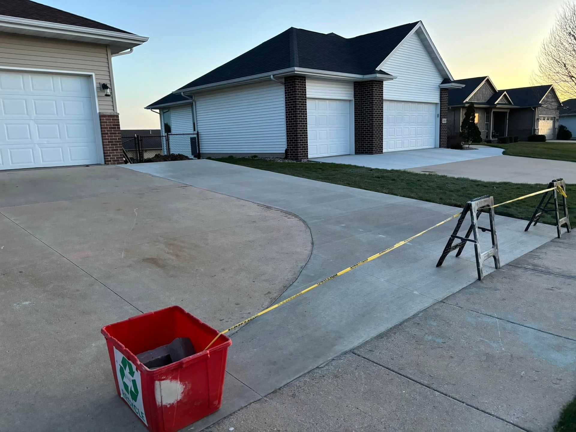 A red recycling bin is attached to caution tape stretched across a sidewalk in front of houses.