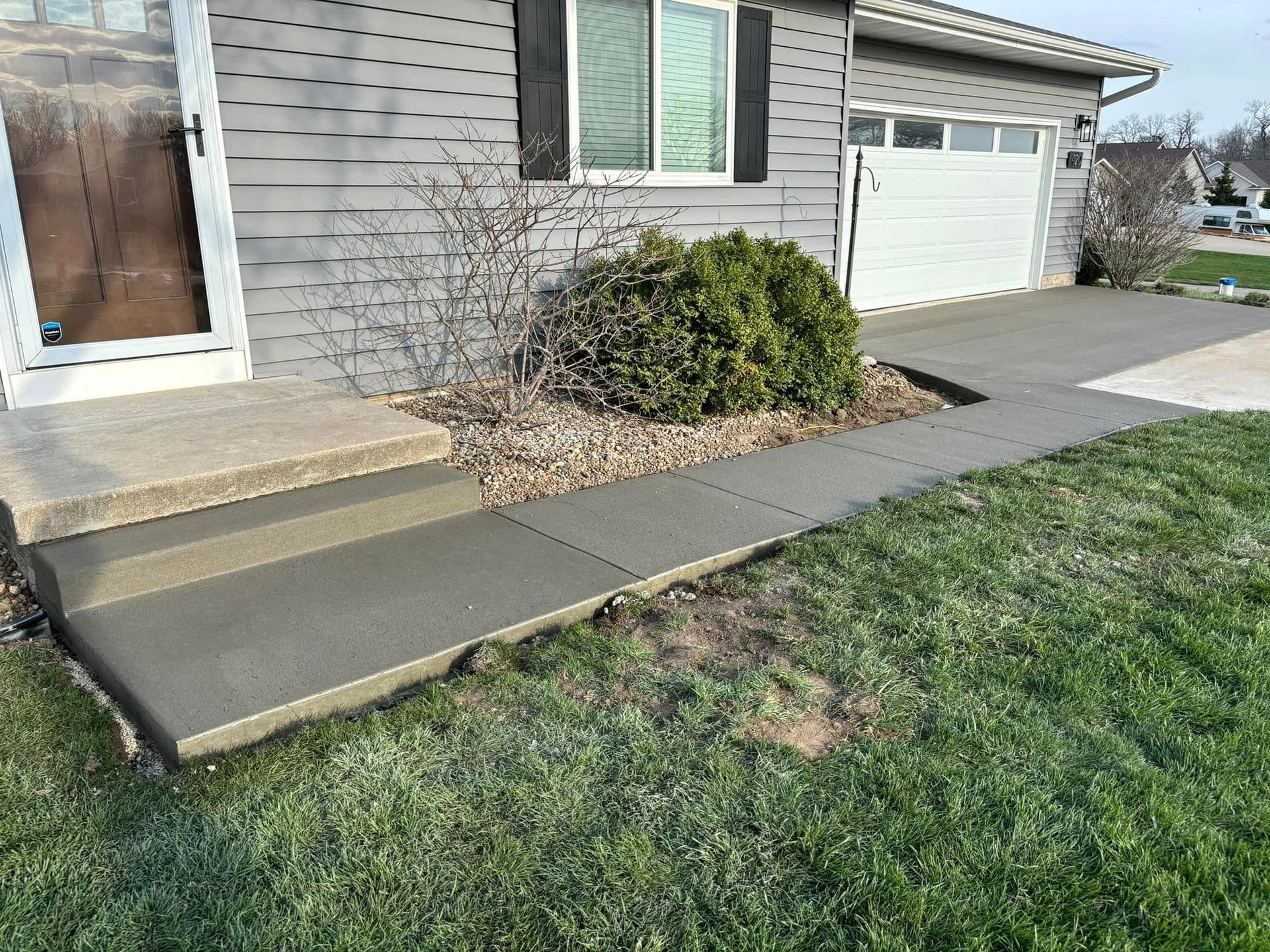 New concrete sidewalk and steps leading to a gray house with a white garage door. Green grass surrounds.