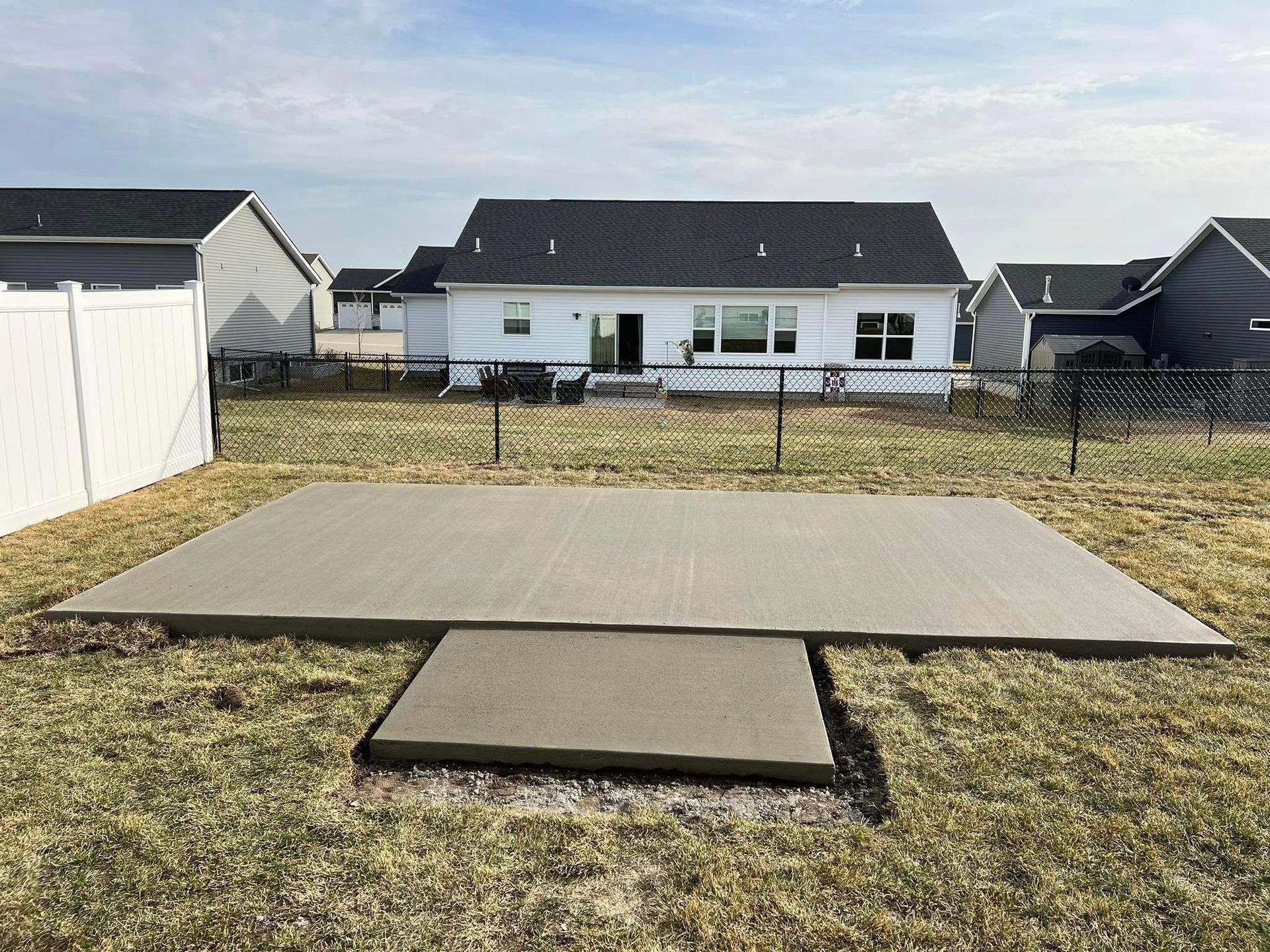 Concrete patio with small pad, in a grassy backyard. Houses and fence are in the background.