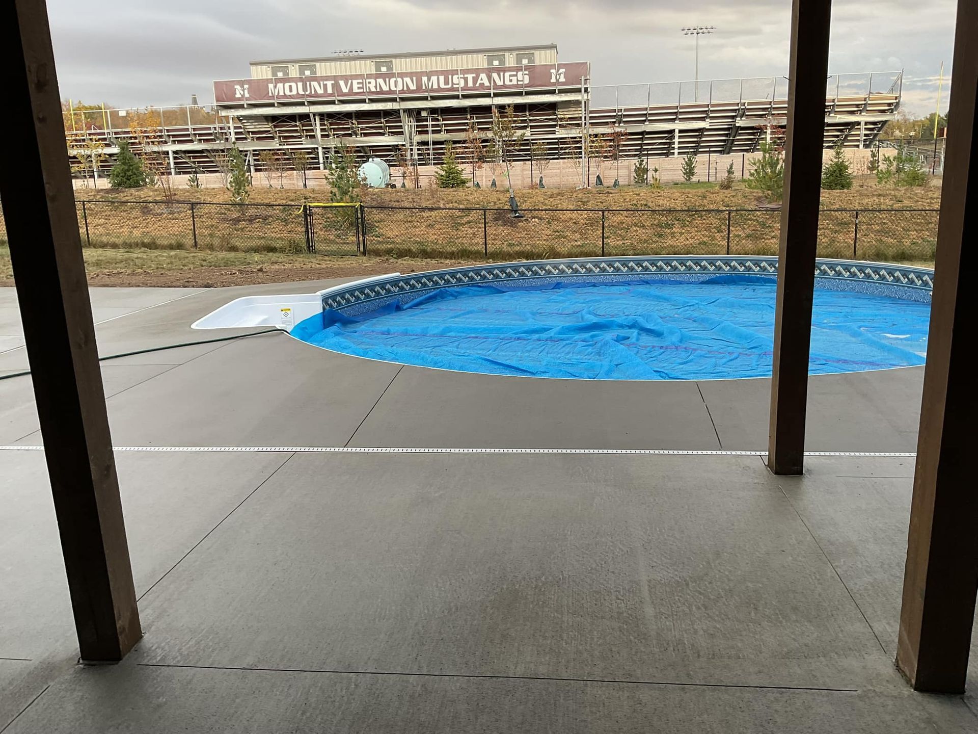 Pool covered with blue tarp, behind a patio with stadium in background. Overcast sky.