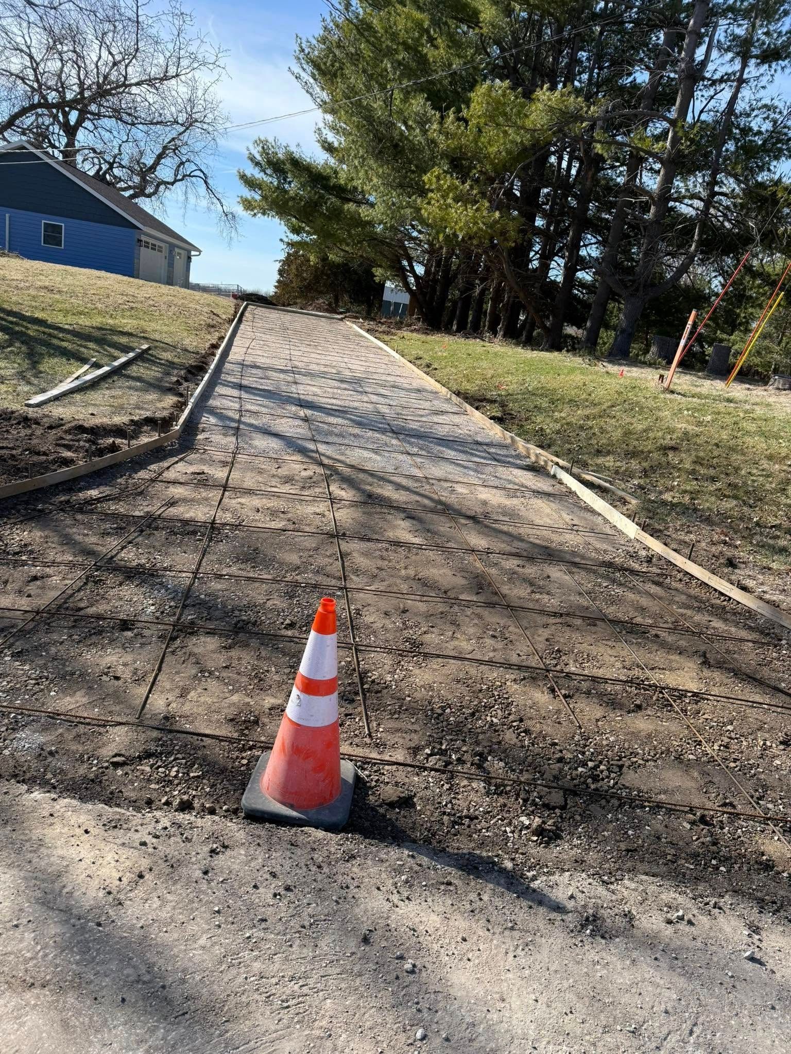 Concrete pathway under construction with safety cone, sloping uphill towards trees.