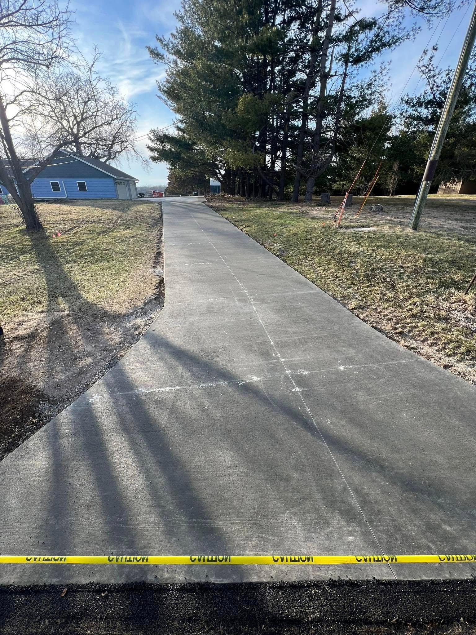 Newly paved asphalt pathway with yellow caution tape, leading towards trees and a blue house.