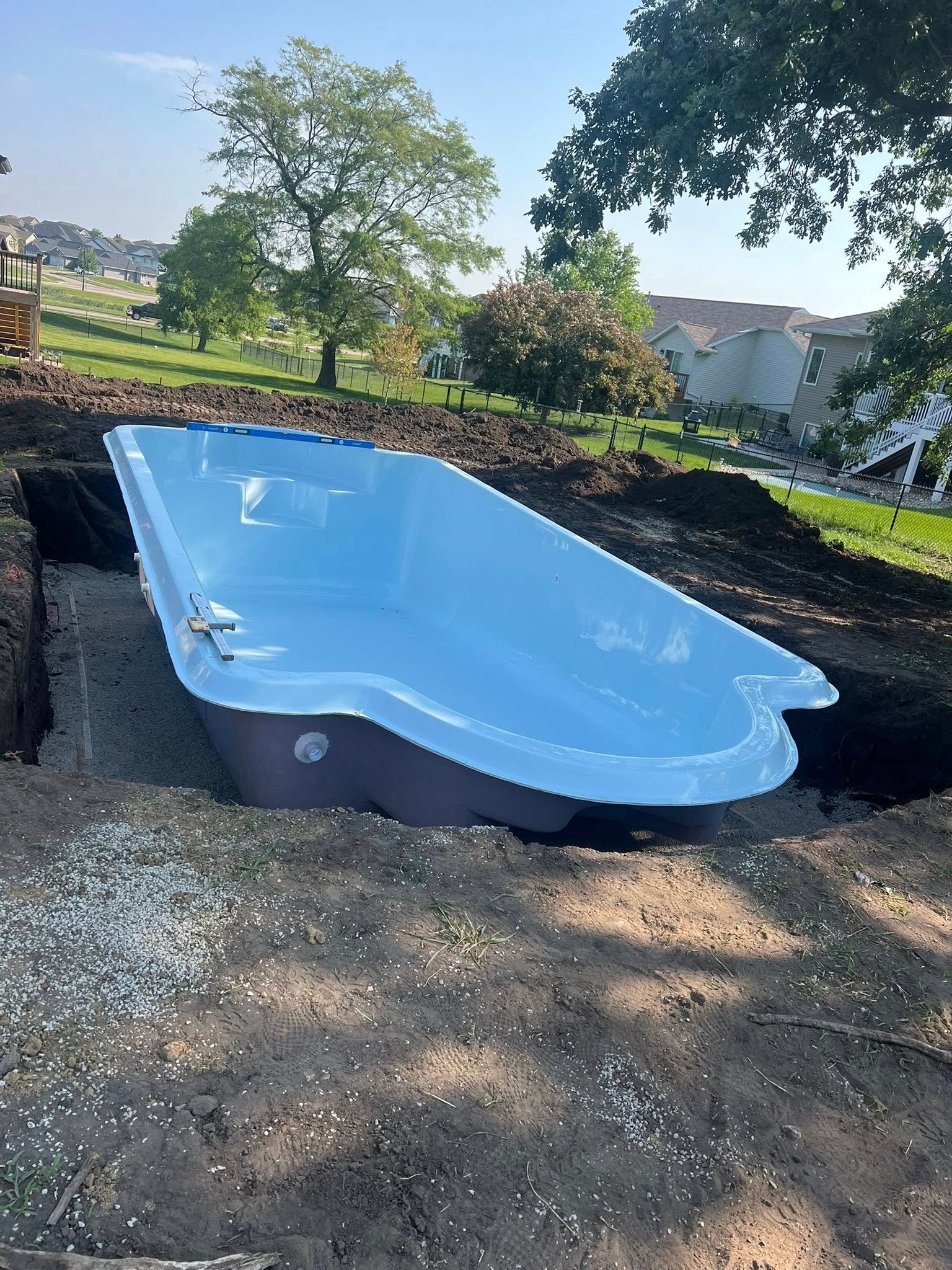 A light blue, empty swimming pool sits in a dirt excavation with a green grassy background.