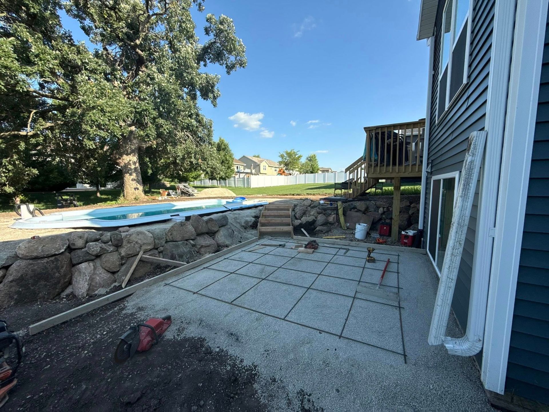 Backyard construction site; concrete patio with rebar, next to a house with a deck. Blue sky.
