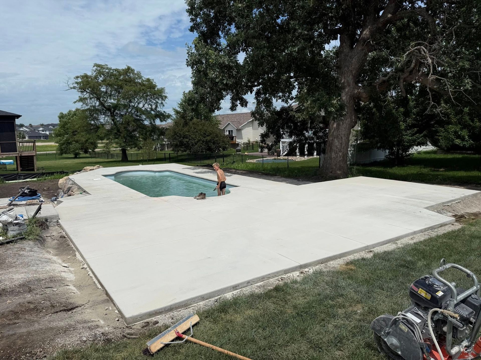 Pool deck construction with a person in the pool. Green grass, large tree, and houses in the background.