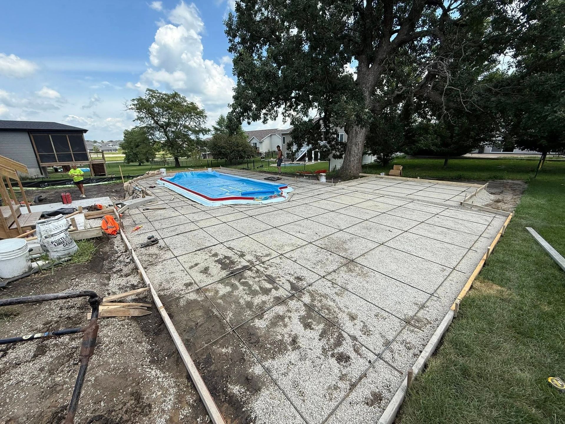 Construction site with a concrete patio being poured around a swimming pool covered in blue tarp.