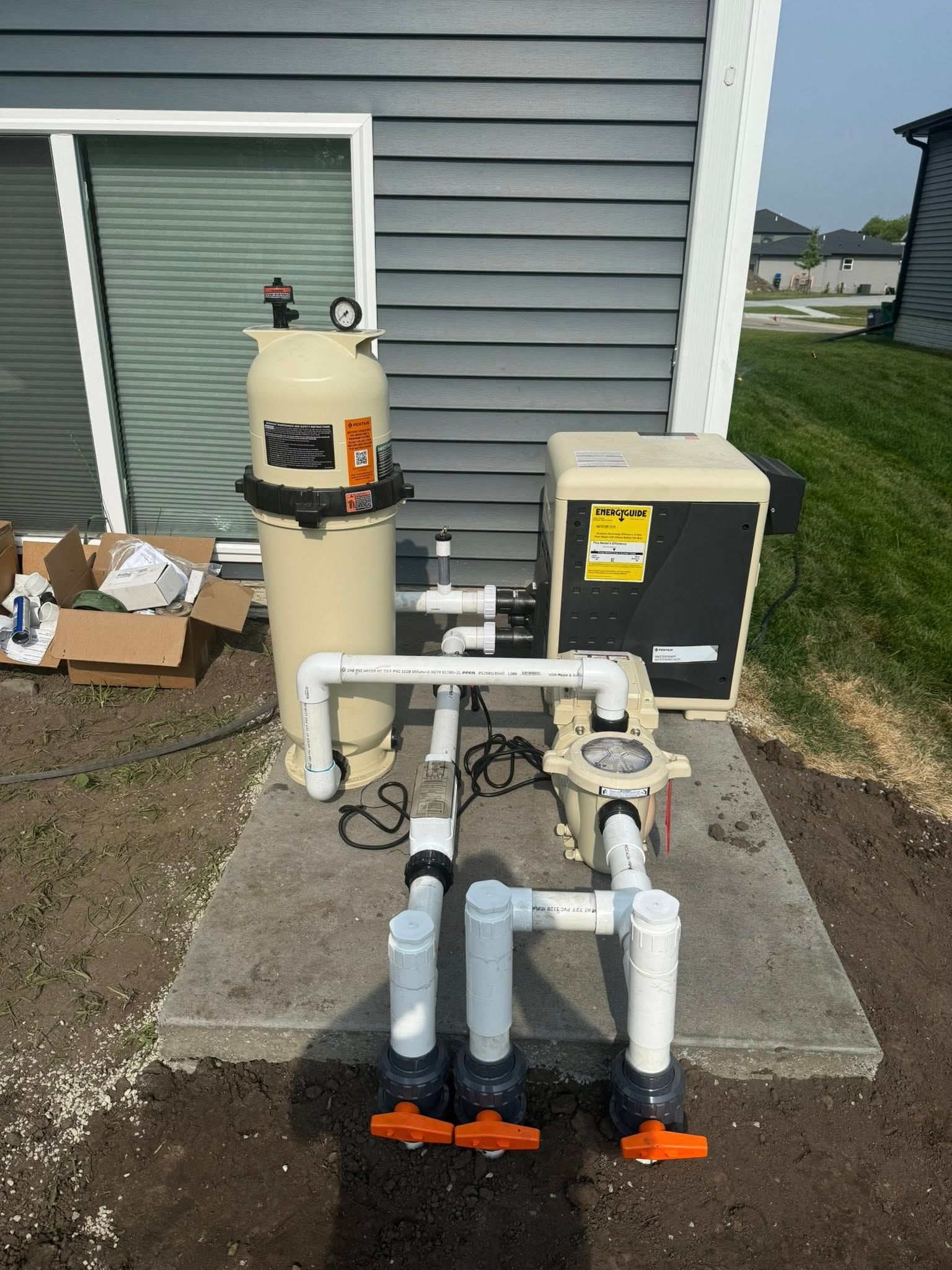 Pool equipment on a concrete pad: filter, pump, heater, and plumbing with three orange valves, next to a building.