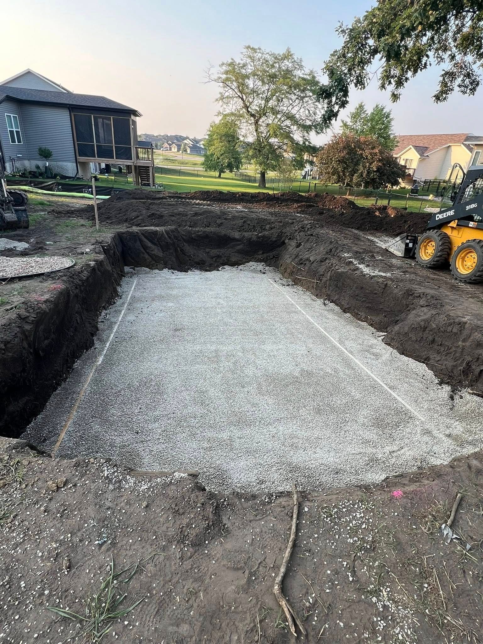 A rectangular excavation with gravel, likely a foundation. Construction equipment and a house in the background.