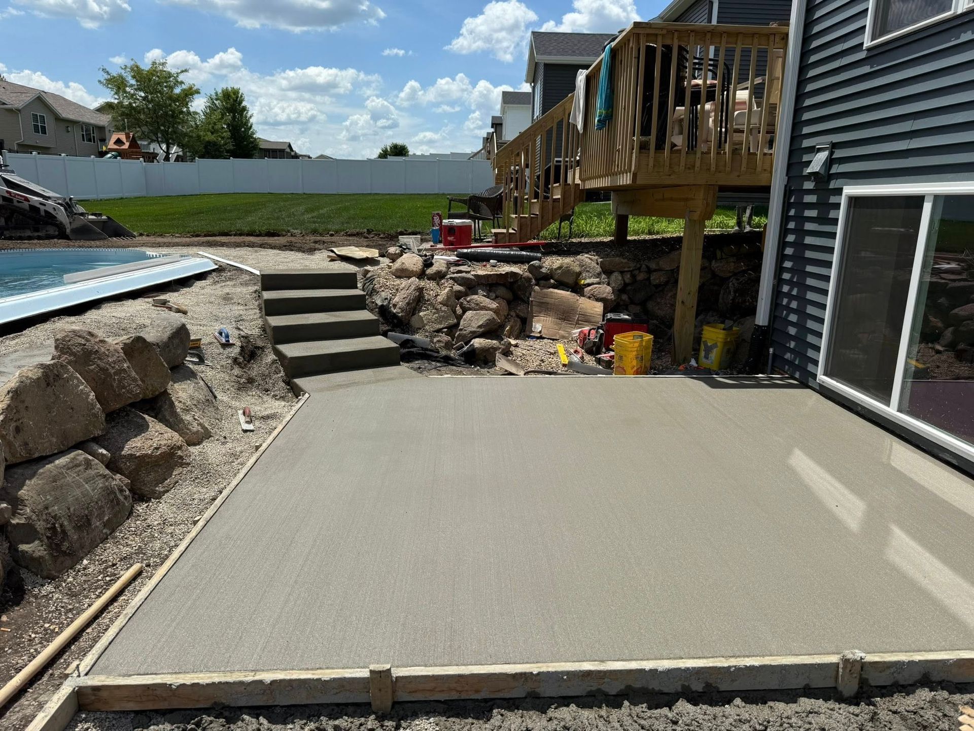 Freshly poured concrete patio next to a house and deck, with a pool and steps in the background.