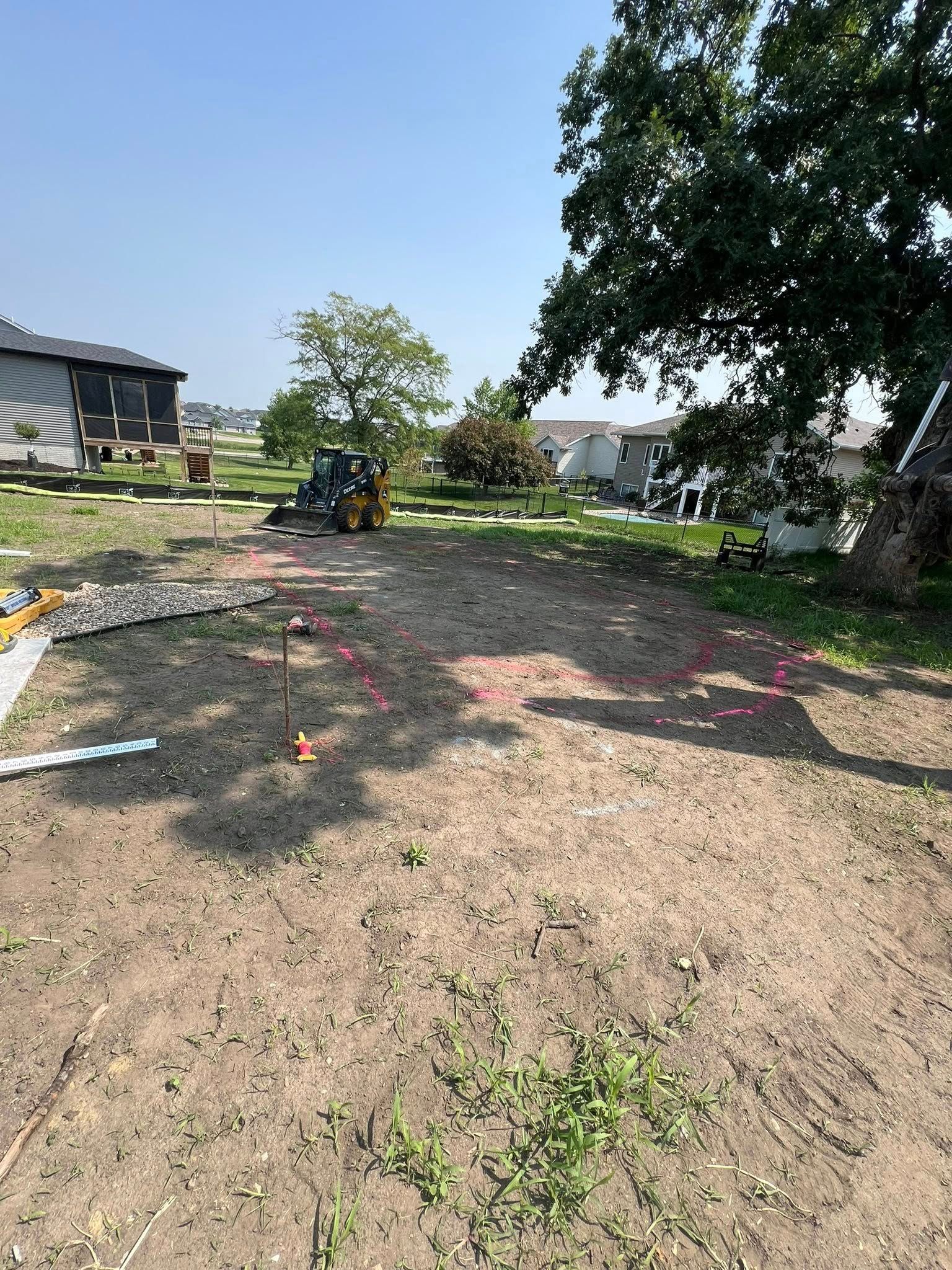Dirt yard marked with pink flags. A small construction vehicle and trees are visible in the background.
