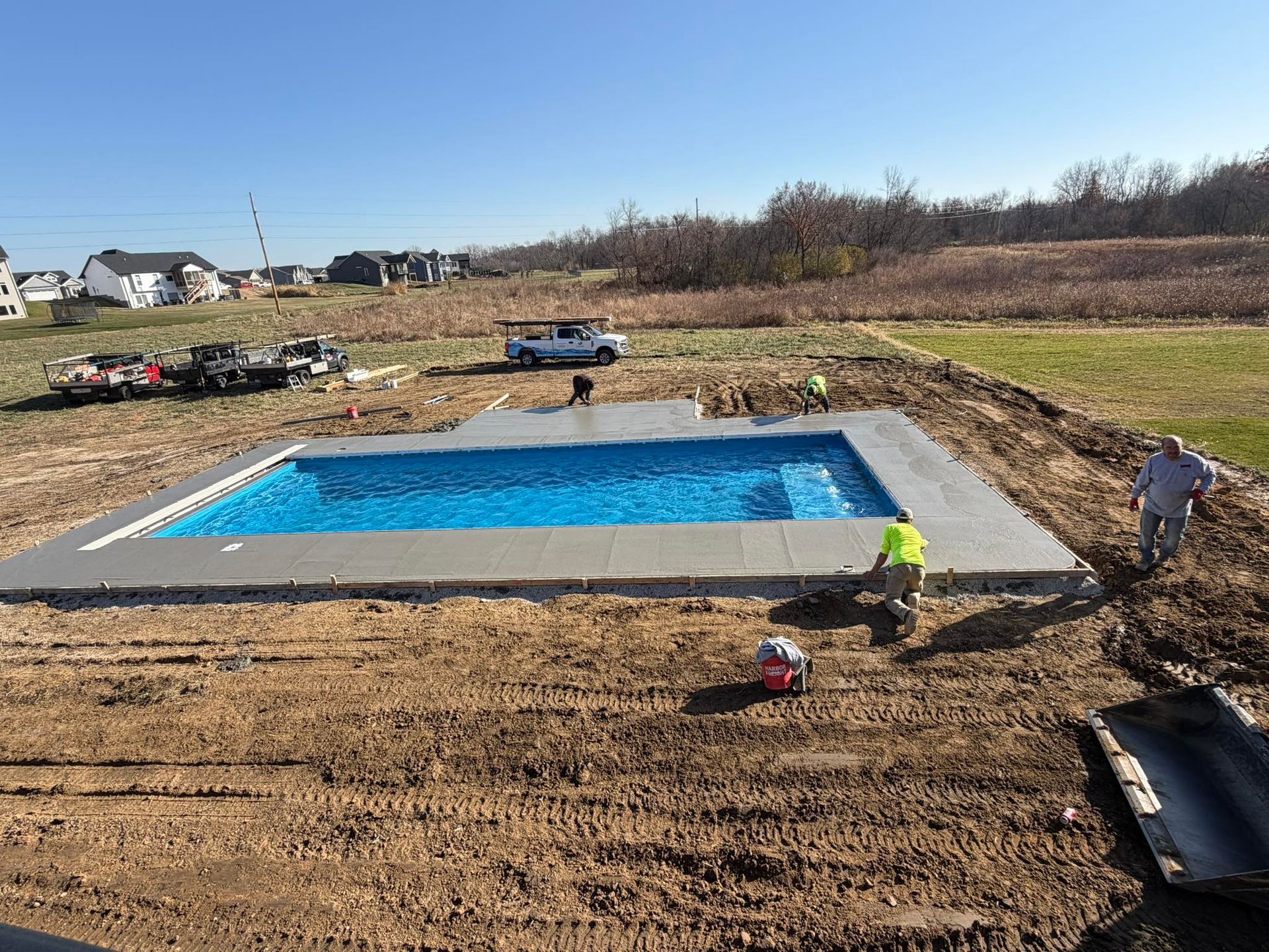 Rectangular swimming pool under construction, surrounded by concrete. Workers in the dirt.