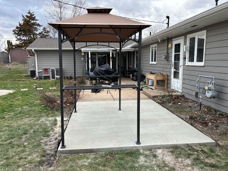 Brown gazebo over a concrete patio in a backyard setting near a gray house.
