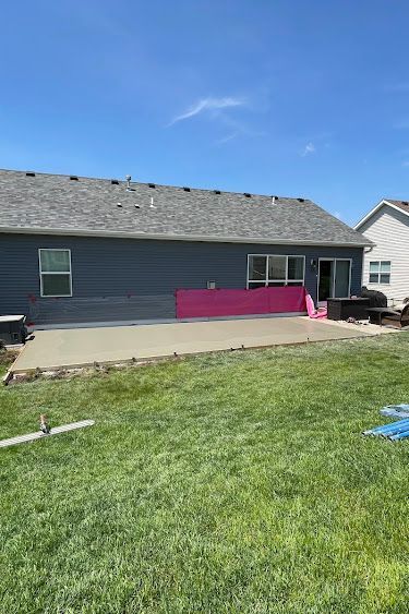 Backyard with gray house and new concrete patio, partially covered with pink material. Blue sky.