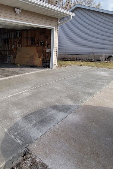 Driveway leading to a garage. Gray concrete with salt residue, next to a gray house.