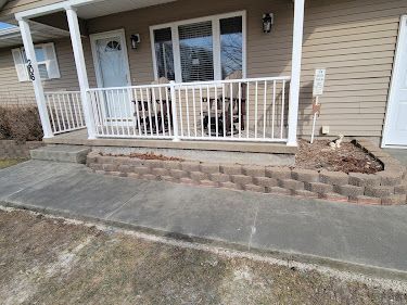 Exterior view of a house with a concrete porch and retaining wall. White railing on porch. Tan siding.