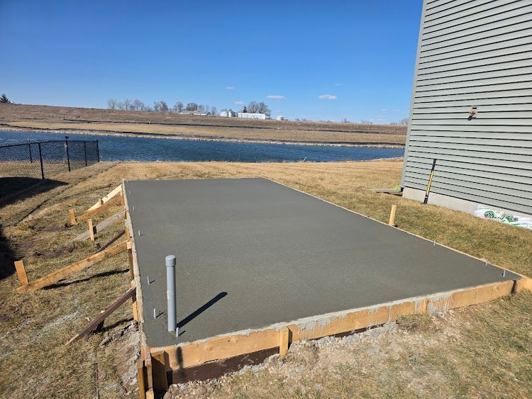 Freshly poured concrete foundation with framing, next to a building and body of water, on a sunny day.