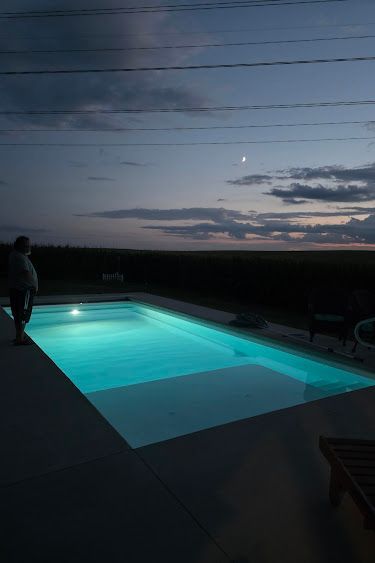 Person standing near a glowing, rectangular pool at dusk; dark sky with moon.
