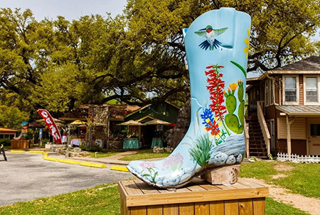 Painted cowboy boot sculpture with fish and flowers on a wooden pedestal outdoors near houses.